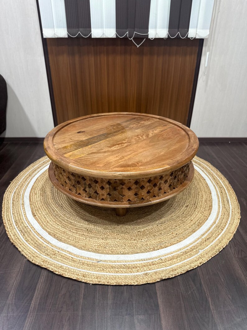May include: A round wooden coffee table with intricate carvings around the base. The table is sitting on a natural fiber rug with white trim.