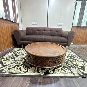 May include: A dark brown wooden coffee table with a carved design around the bottom edge. The table is sitting on a black and green patterned rug in front of a brown sofa.