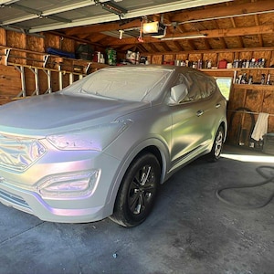 May include: A silver SUV is parked inside a garage. The vehicle is covered in a protective wrap, with the front end and hood visible. The car has black wheels and is surrounded by tools and equipment.