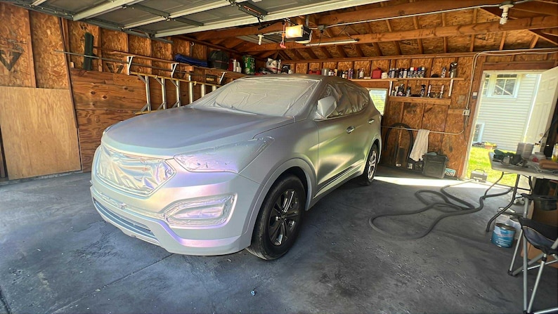 May include: A silver SUV is parked inside a garage. The vehicle is covered in a protective wrap, with the front end and hood visible. The car has black wheels and is surrounded by tools and equipment.