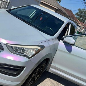 May include: A light gray SUV with a pearlescent finish, parked on a residential street. The car's door is open, revealing the interior. The vehicle has black wheels and a chrome grill.