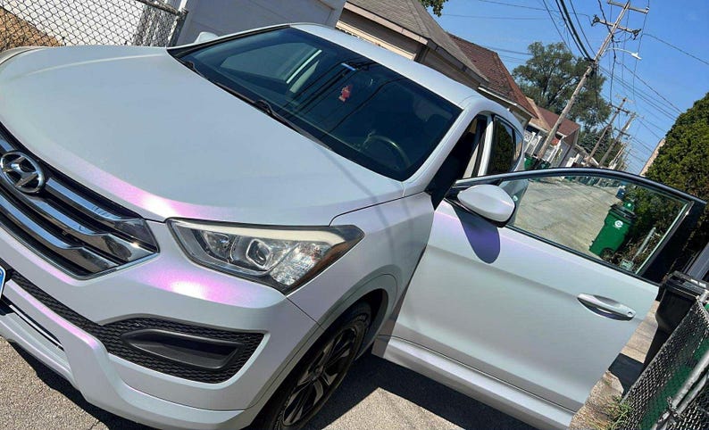 May include: A light gray SUV with a pearlescent finish, parked on a residential street. The car's door is open, revealing the interior. The vehicle has black wheels and a chrome grill.