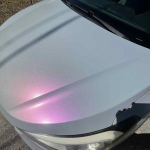 May include: A car hood with a pearlescent finish, transitioning from a light blue-gray to a vibrant pink and purple hue. The hood's surface reflects light, creating a dynamic color shift. The car's headlight is visible.