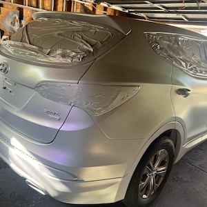 May include: A silver Hyundai Santa Fe Sport SUV, partially masked for painting. The vehicle is in a garage, with the rear and side panels visible. The car's wheels are black, and the paint job appears to be in progress.