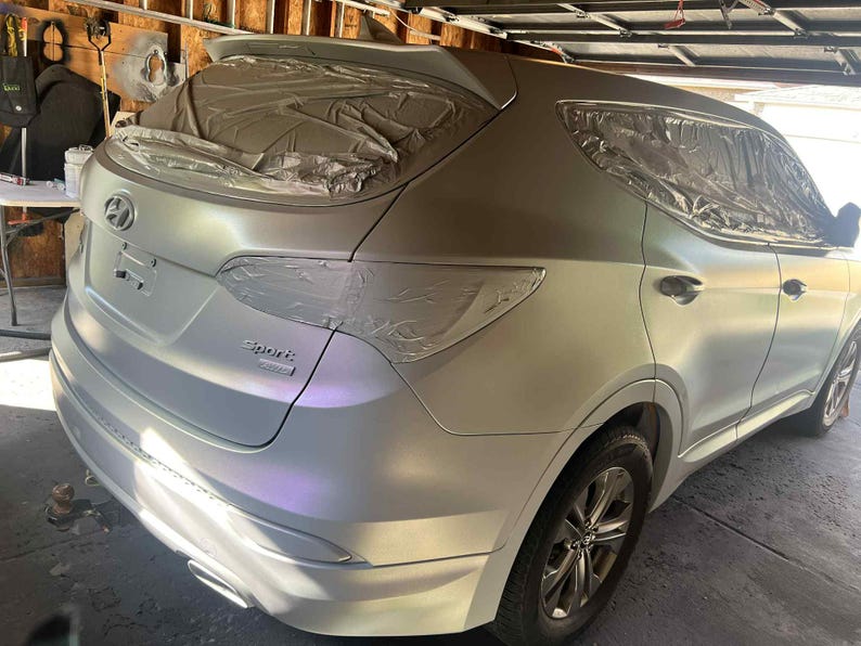 May include: A silver Hyundai Santa Fe Sport SUV, partially masked for painting. The vehicle is in a garage, with the rear and side panels visible. The car's wheels are black, and the paint job appears to be in progress.