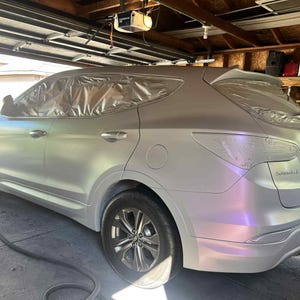 May include: A silver SUV with sections covered in plastic sheeting, likely for painting. The vehicle has a pearlescent finish that shifts colors in the light. The wheels are silver, and the car is parked in a garage.