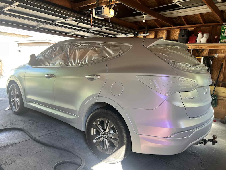 May include: A silver SUV with sections covered in plastic sheeting, likely for painting. The vehicle has a pearlescent finish that shifts colors in the light. The wheels are silver, and the car is parked in a garage.