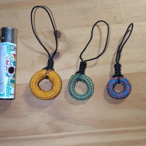 Pode incluir: Três pequenos chaveiros coloridos com um design circular são exibidos em uma superfície de madeira. Os chaveiros são em ouro, verde e vermelho/azul. Um isqueiro com um design floral está à esquerda.