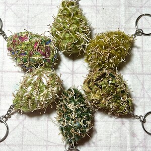 Pode incluir: Seis chaveiros artesanais em forma de estrela. Cada chaveiro apresenta um design fofo e texturizado em tons de verde, branco e dourado, preso a um porta-chaves prateado. Os chaveiros estão dispostos em um padrão de estrela.