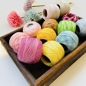 May include: A wooden box filled with colorful balls of yarn. The yarn is in shades of pink, red, yellow, green, blue, and white. The yarn is arranged in a circular pattern.