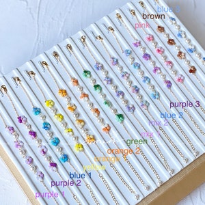 May include: A display of delicate flower charm bracelets with pearl accents in various colors, including blue, purple, orange, green, brown, and pink. The bracelets are arranged in rows on a white surface with labels indicating the color of each bracelet.