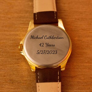 Puede incluir: Primer plano de un reloj dorado con correa de cuero marr&oacute;n. La parte trasera del reloj est&aacute; grabada con el nombre "Michael Cuthbertson", el texto "42 Years" y la fecha "5/27/2023". El reloj est&aacute; sobre un fondo de madera.