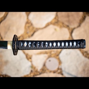 Puede incluir: Primer plano del mango de una espada katana. El mango est&aacute; envuelto en un cord&oacute;n negro con un patr&oacute;n de diamantes. La guarda es negra y circular. La hoja es parcialmente visible, con un acabado verde oscuro y dorado.