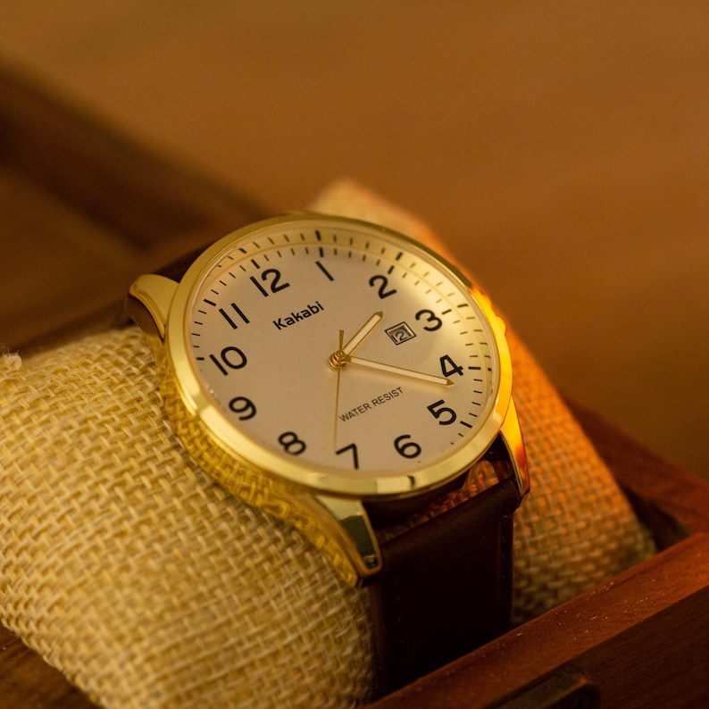 Puede incluir: Un reloj de pulsera Kakabi dorado con esfera crema, n&uacute;meros negros y correa de cuero marr&oacute;n. El reloj se exhibe sobre una superficie de arpillera beige. La esfera indica "Water Resist".