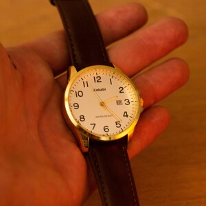Puede incluir: Un reloj de pulsera dorado con esfera blanca y n&uacute;meros negros. El reloj tiene una correa de cuero marr&oacute;n. La esfera muestra la marca "Kakabi" y las palabras "Water Resist".