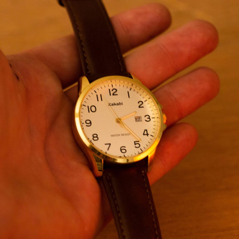Puede incluir: Un reloj de pulsera dorado con esfera blanca y n&uacute;meros negros. El reloj tiene una correa de cuero marr&oacute;n. La esfera muestra la marca "Kakabi" y las palabras "Water Resist".