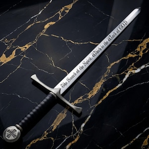 May include: A silver-toned metal sword with a black leather-wrapped handle and a circular pommel featuring a cross emblem. The blade is engraved with the black text, The Sword of the Spirit, Which is the Word of GOD, in a gothic script font.