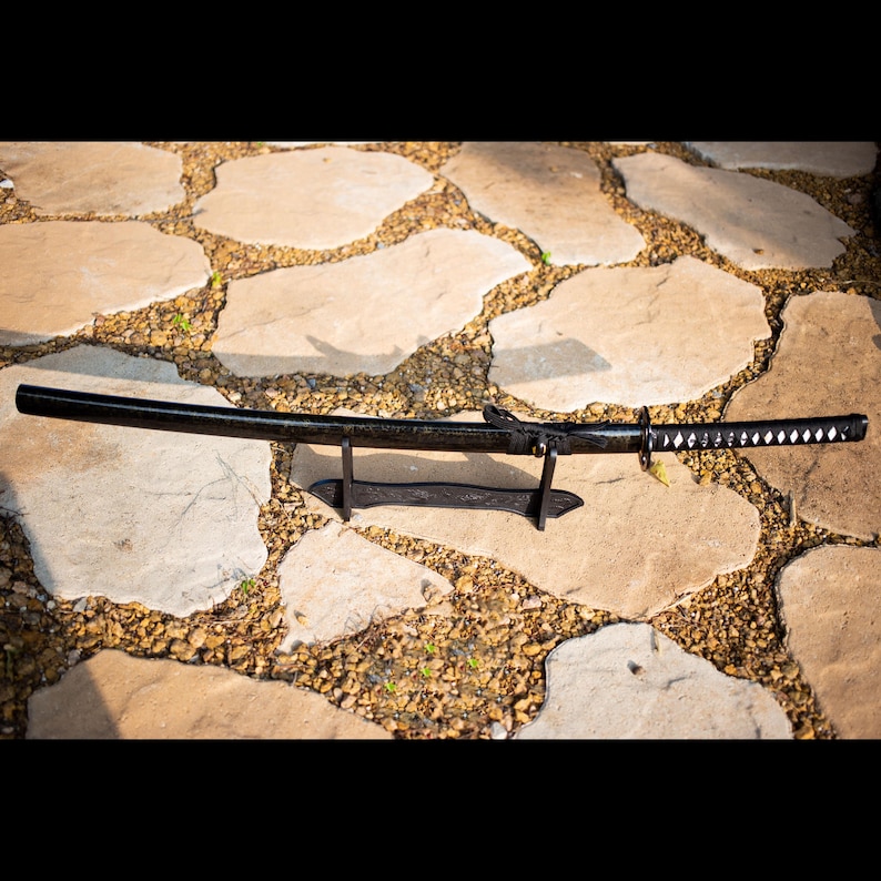 Puede incluir: Una espada katana negra con una hoja curva, apoyada sobre un soporte negro. El mango de la espada tiene un patr&oacute;n blanco y negro. El fondo es un pavimento de piedra.
