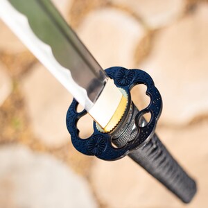Puede incluir: Primer plano de una katana japonesa. La hoja plateada tiene un borde ondulado. La guarda es azul oscuro con un dise&ntilde;o floral y un mango negro. La espada se encuentra sobre un fondo neutro borroso.
