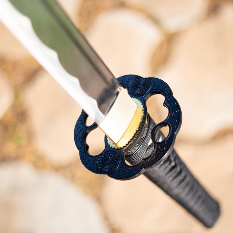 Puede incluir: Primer plano de una katana japonesa. La hoja plateada tiene un borde ondulado. La guarda es azul oscuro con un dise&ntilde;o floral y un mango negro. La espada se encuentra sobre un fondo neutro borroso.