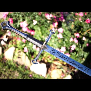 May include: A silver sword with a black handle and a blade engraved with the text "Sir Michael Cuthbertson II." The sword has a decorative hilt and crossguard. The background features green foliage and pink flowers.
