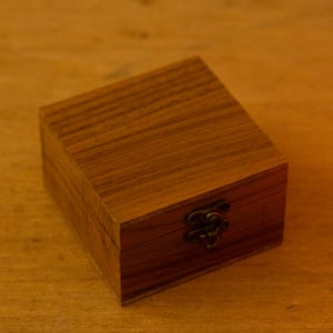 Puede incluir: Peque&ntilde;a caja cuadrada de madera clara con tapa con bisagras y cierre met&aacute;lico. La madera tiene un veteado visible. El cierre es de color bronce oscuro.