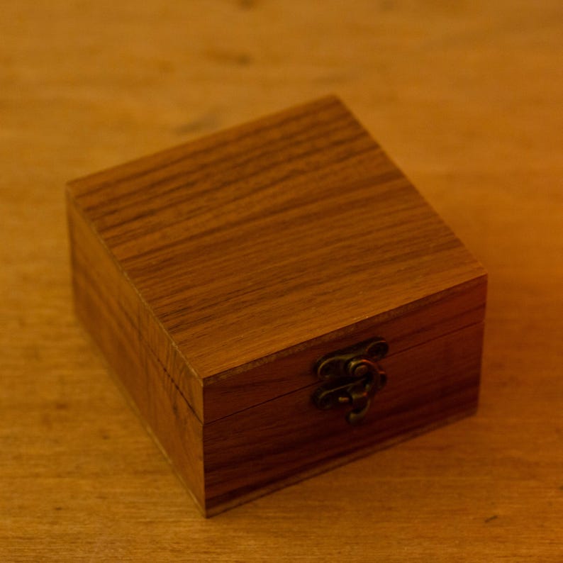 Puede incluir: Peque&ntilde;a caja cuadrada de madera clara con tapa con bisagras y cierre met&aacute;lico. La madera tiene un veteado visible. El cierre es de color bronce oscuro.