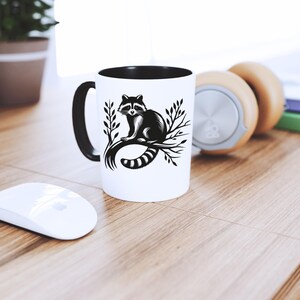 May include: White ceramic mug with a black interior and handle. The mug features a black and white illustration of a raccoon sitting on a branch. The mug is on a wooden surface, next to a white mouse and headphones.