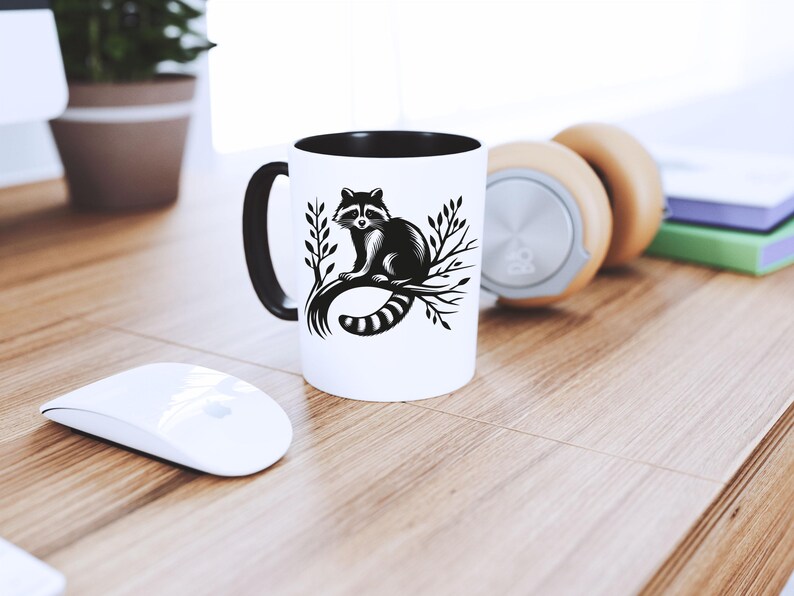 May include: White ceramic mug with a black interior and handle. The mug features a black and white illustration of a raccoon sitting on a branch. The mug is on a wooden surface, next to a white mouse and headphones.