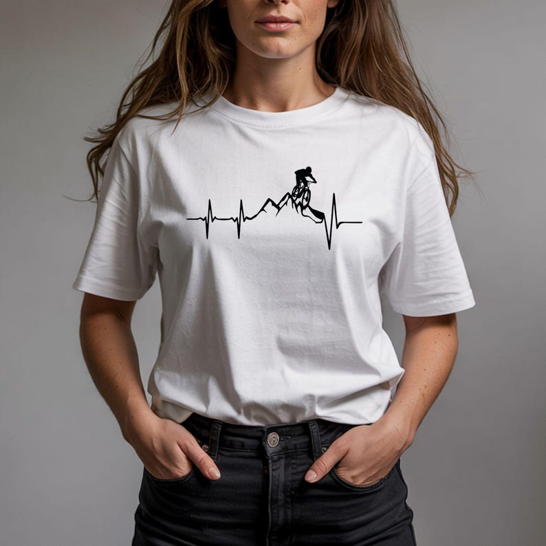 May include: White t-shirt featuring a black graphic of a mountain biker on a mountain range, integrated into a heartbeat line design. The shirt is a classic crew neck style.