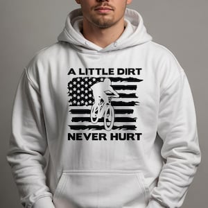 May include: White hooded sweatshirt with a black graphic of a mountain biker and an American flag design. The text reads "A LITTLE DIRT NEVER HURT". The sweatshirt has a front pocket and a drawstring hood.