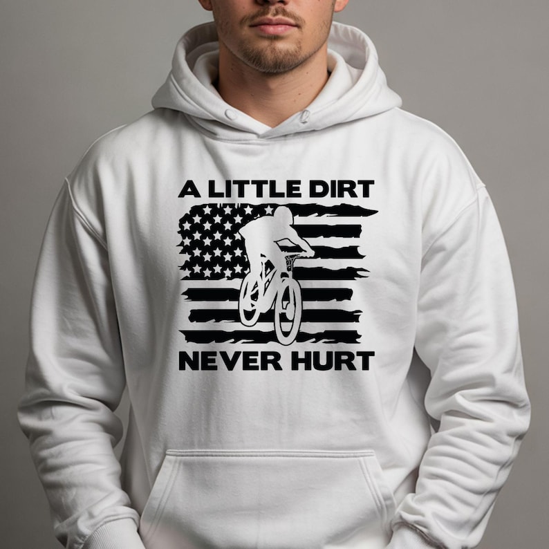 May include: White hooded sweatshirt with a black graphic of a mountain biker and an American flag design. The text reads "A LITTLE DIRT NEVER HURT". The sweatshirt has a front pocket and a drawstring hood.