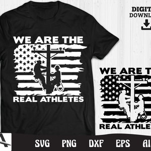 Puede incluir: Una camiseta negra con un diseño blanco que presenta dos figuras de silueta de electricistas trabajando en una línea eléctrica frente a una bandera estadounidense. El texto "We are the real athletes" está impreso debajo del diseño.