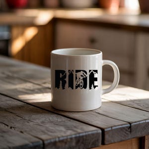 May include: White ceramic mug with the word "RIDE" in black block letters. The letter "I" features a silhouette of a mountain biker. The mug is on a rustic wooden table.