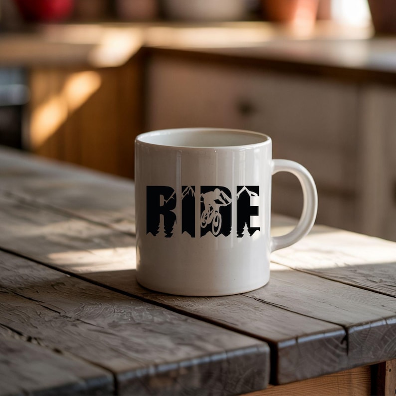 May include: White ceramic mug with the word "RIDE" in black block letters. The letter "I" features a silhouette of a mountain biker. The mug is on a rustic wooden table.
