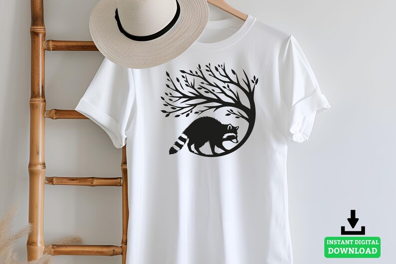 May include: White t-shirt featuring a black graphic of a raccoon inside a tree. The raccoon has a striped tail and is surrounded by tree branches. A straw hat hangs on a wooden ladder. The words "INSTANT DIGITAL DOWNLOAD" are in a green box.