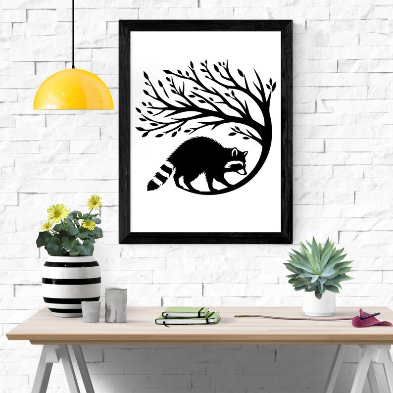 May include: Black and white framed art print featuring a raccoon silhouette under a tree. The artwork is displayed on a white brick wall, with a yellow pendant light and a wooden table with decorative items in the foreground.