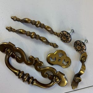May include: A collection of antique brass drawer pulls and knobs. The set includes two long pulls, two small knobs, a decorative escutcheon, and a drop pull. The hardware features ornate detailing and a warm, aged finish, perfect for vintage furniture restoration projects.