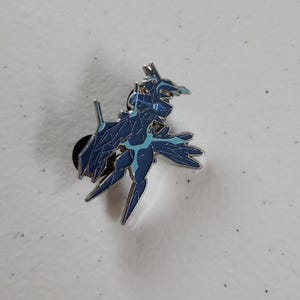 May include: A dark blue and light blue enamel pin featuring a stylized creature design. The pin has a silver outline and a black pin back. The creature has sharp angles and a detailed design, with light blue accents.