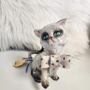 Può includere: Statuina di gatto in ceramica con corpo bianco e grigio, occhi verdi e papillon a pois. Il gatto ha un'espressione stravagante, leggermente triste. Una piccola etichetta di legno è attaccata alla coda con un nastro blu.