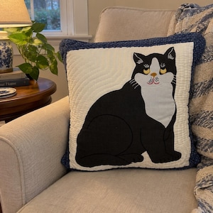May include: A square throw cushion featuring a quilted off-white background with a black and white cat appliqué. The cat has yellow eyes, a pink nose, and a white chest. The cushion is finished with a dark blue patterned border and wavy stitching.