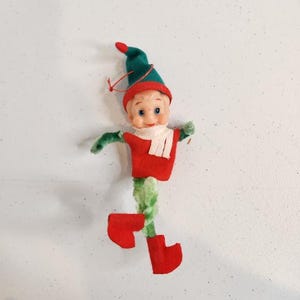 May include: A vintage Christmas elf doll. The doll has a green hat, red boots, and a red shirt. The elf has a white scarf and green arms. The legs are made of a fuzzy green material. The doll's face is a light skin tone with blue eyes and a smiling expression.