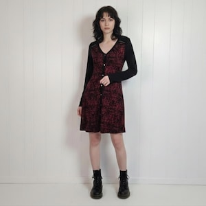 May include: A patterned button-down dress in black and burgundy. The dress features long black sleeves and a V-neck. The model is wearing black boots.