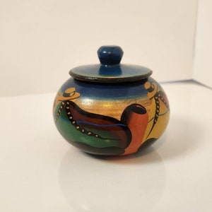 以下が含まれることがあります： 青い蓋が付いた小さな丸い木製の壺。壺には、帽子をかぶった伝統的な服装の人々が描かれたカラフルな絵が描かれています。壺は、ジュエリーや小物などの小さなものを保管するのに最適です。
