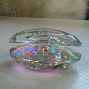 May include: A clear, faceted glass clam shell with an iridescent interior. A faux pearl sits inside the bottom half of the shell. The top half of the shell is open, revealing the pearl. The shell is on a white surface.