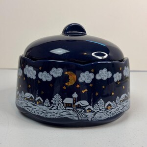 Op de afbeelding: Een donkerblauwe keramische pot met deksel, voorzien van een handgeschilderd winterdorp-tafereel. Het ontwerp toont witte, besneeuwde huizen, groenblijvende bomen en grijze wolken tegen een nachtelijke hemel vol gouden sterren en een maansikkel.