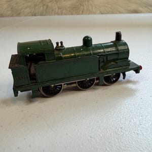May include: A dark green toy steam locomotive featuring black wheels and the number 3337 printed on the side of the cab. The metal train model has a chimney, a dome, and a small red detail on the rear buffer, showing signs of paint wear.