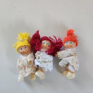 Könnte beinhalten: Drei Vintage-Puppenfiguren mit Garnhaaren in Gelb, Rot und Orange. Die Puppen tragen weiße und gemusterte Kleider und Schuhe. Die Puppen haben blaue Augen und sind in einer spielerischen Anordnung angeordnet.