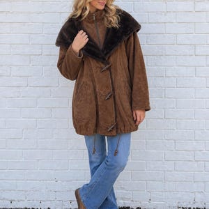 May include: A brown suede coat featuring a large dark brown faux fur collar, a central zipper closure, and three wooden toggle buttons. The coat has long sleeves, side pockets, and a drawstring hem, paired with light blue denim jeans and brown boots.