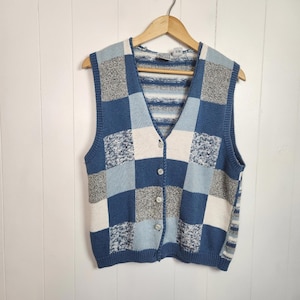 Może przedstawiać: Niebieska, biała i szara kamizelka z dzianiny w stylu patchwork z dekoltem w serek i zapięciem na guziki. Bezrękawnik wisi na drewnianym wieszaku. Wzór obejmuje kwadraty i paski. Tło jest białe.
